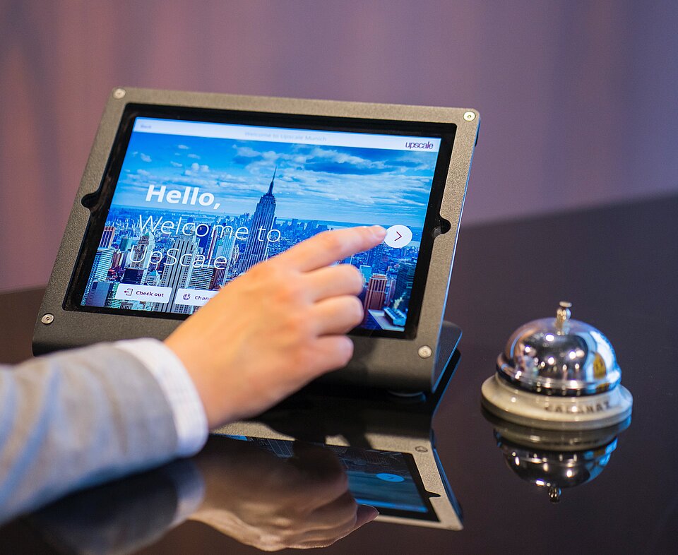 Eine Hand nimmt online, an einem Tablet eine Hotelreservierung vor. Daneben steht eine Hotelklocke. 