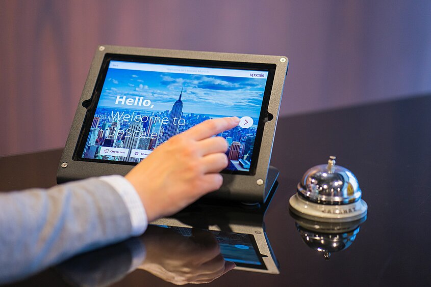 Eine Hand nimmt online, an einem Tablet eine Hotelreservierung vor. Daneben steht eine Hotelklocke. 