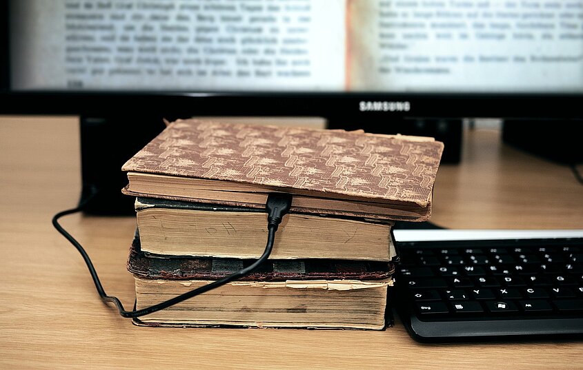 Alte Bücher liegen auf einem Tisch, ein Ladekapbel führt in ein Buch. Daneben liegt eine Tastatur. 