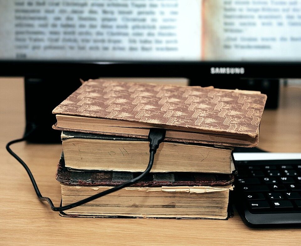 Alte Bücher liegen auf einem Tisch, ein Ladekapbel führt in ein Buch. Daneben liegt eine Tastatur. 